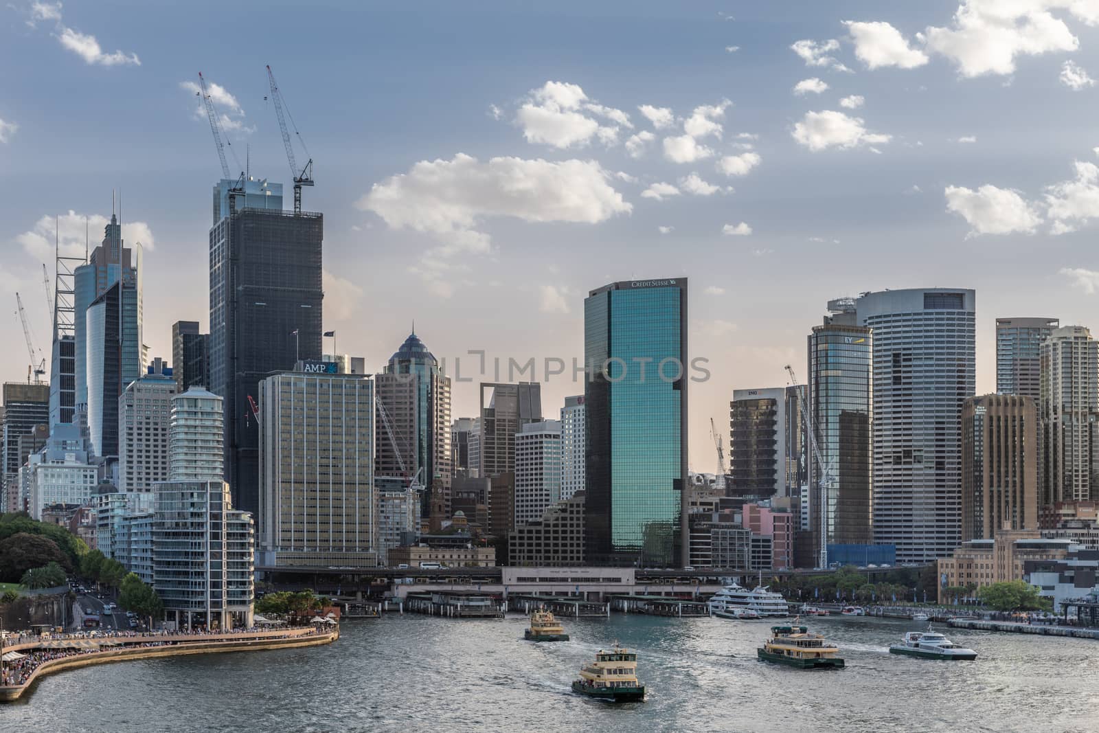 Sydney, Australia - February 12, 2019: Wide shot of Ferry terminal and Circular Quay Railway Station plus skyline in back. Highrises under construction with cranes. Evening shot with light blue sky.