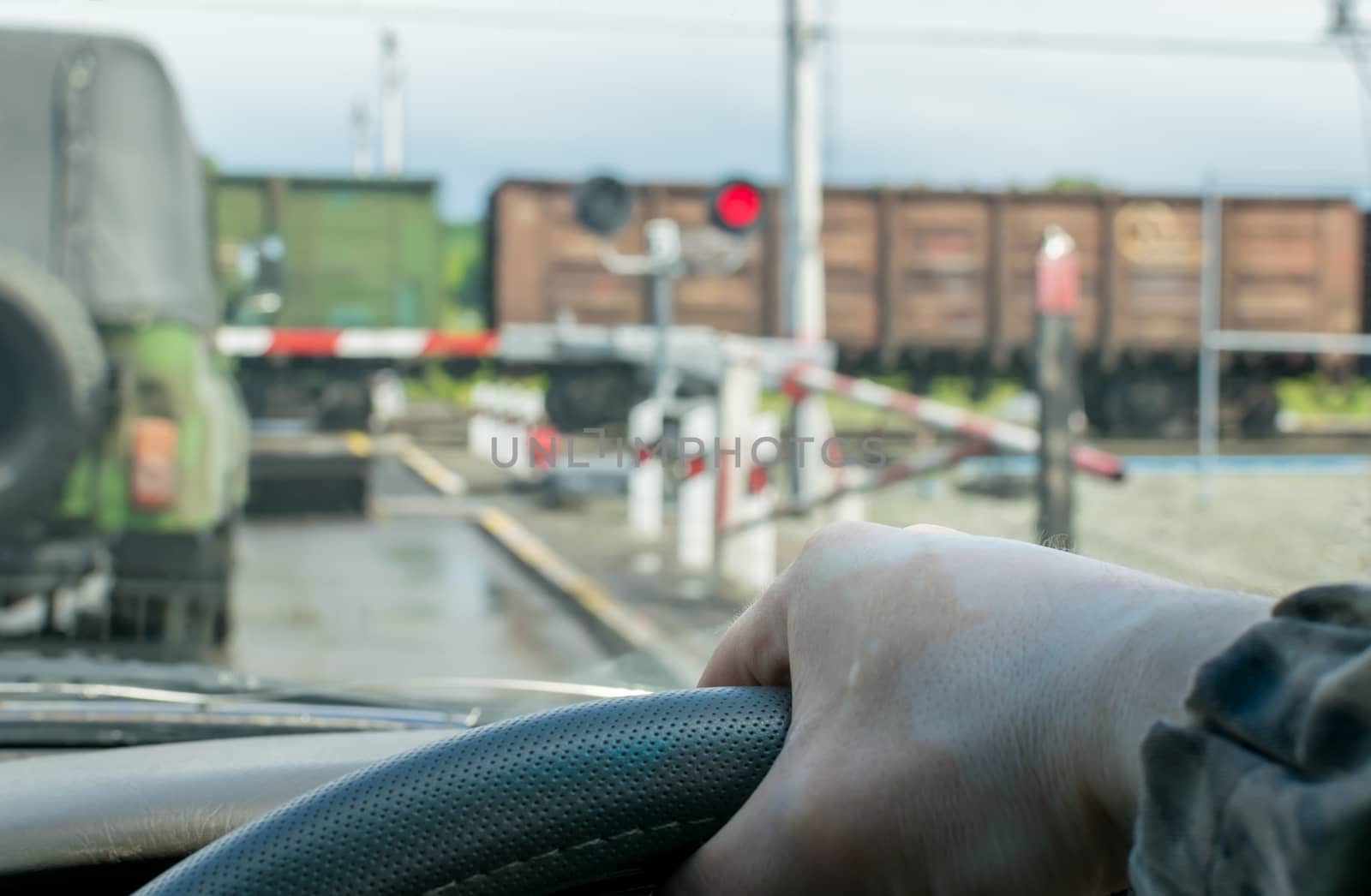 View of the driver hand on the steering wheel of the car, which stopped in front of a closed railway crossing at a red traffic light. The driver waits for the freight train to pass