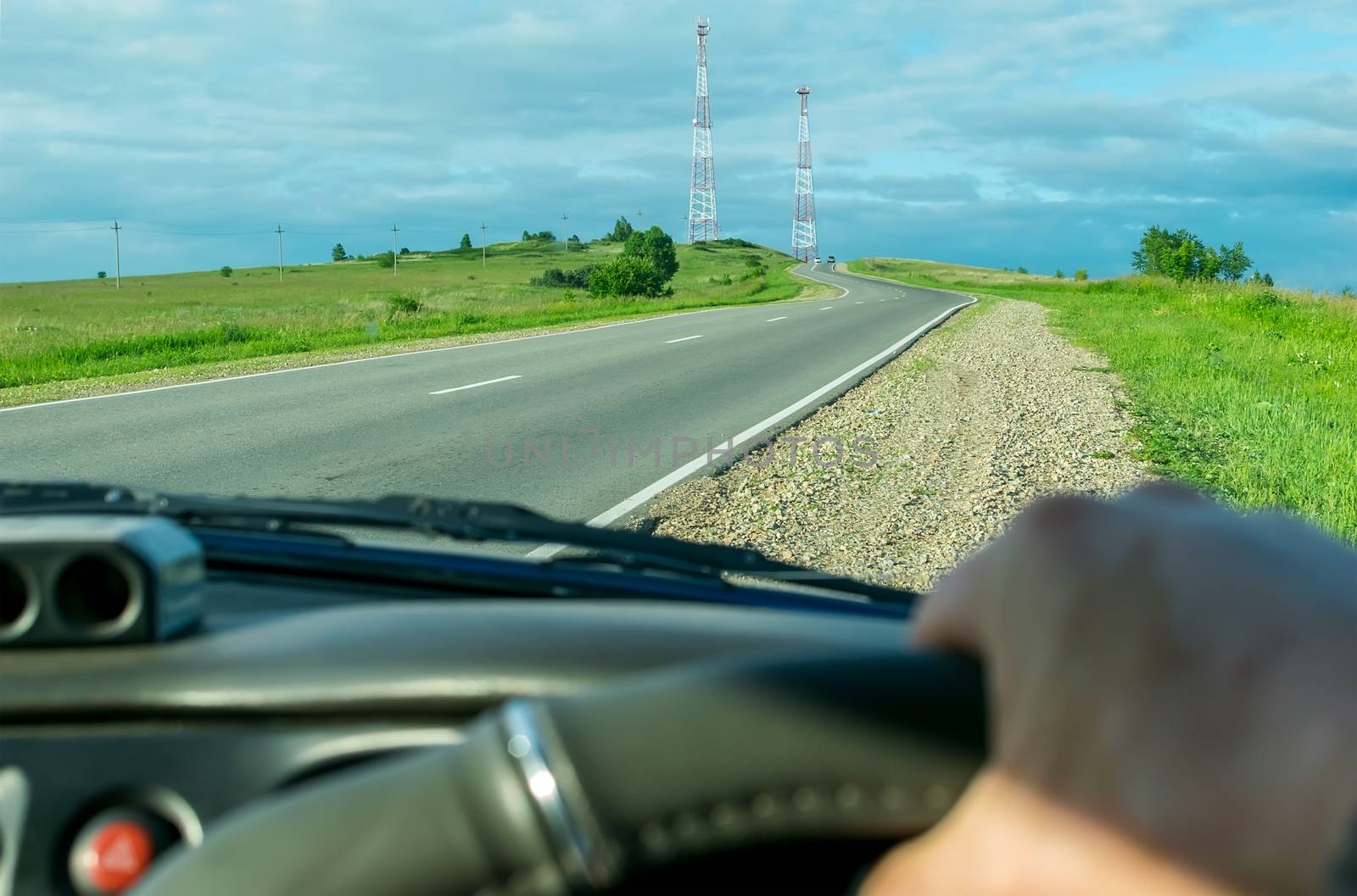 View from the driver's side of the car hands on the steering wheel on the background of the landscape and going up on the hill road with radio towers on the top
