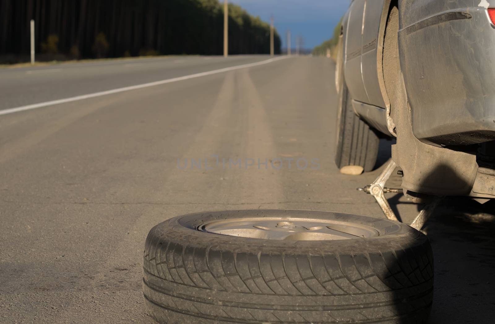 Spare wheel lying near the car. The car is mounted on the Jack. View asphalt road at sunset