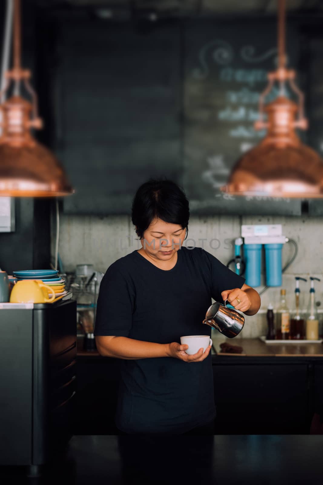 Asian woman 40s white skin work as a barista pouring latte froth to make a coffee latte art into the white coffee cup for serve to customers in the coffee shop cafe