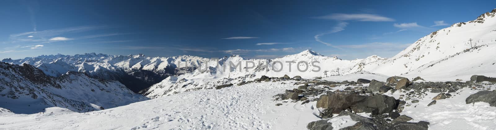 Panoramic view down snow covered valley in alpine mountain range on blue sky background with rock in foreground