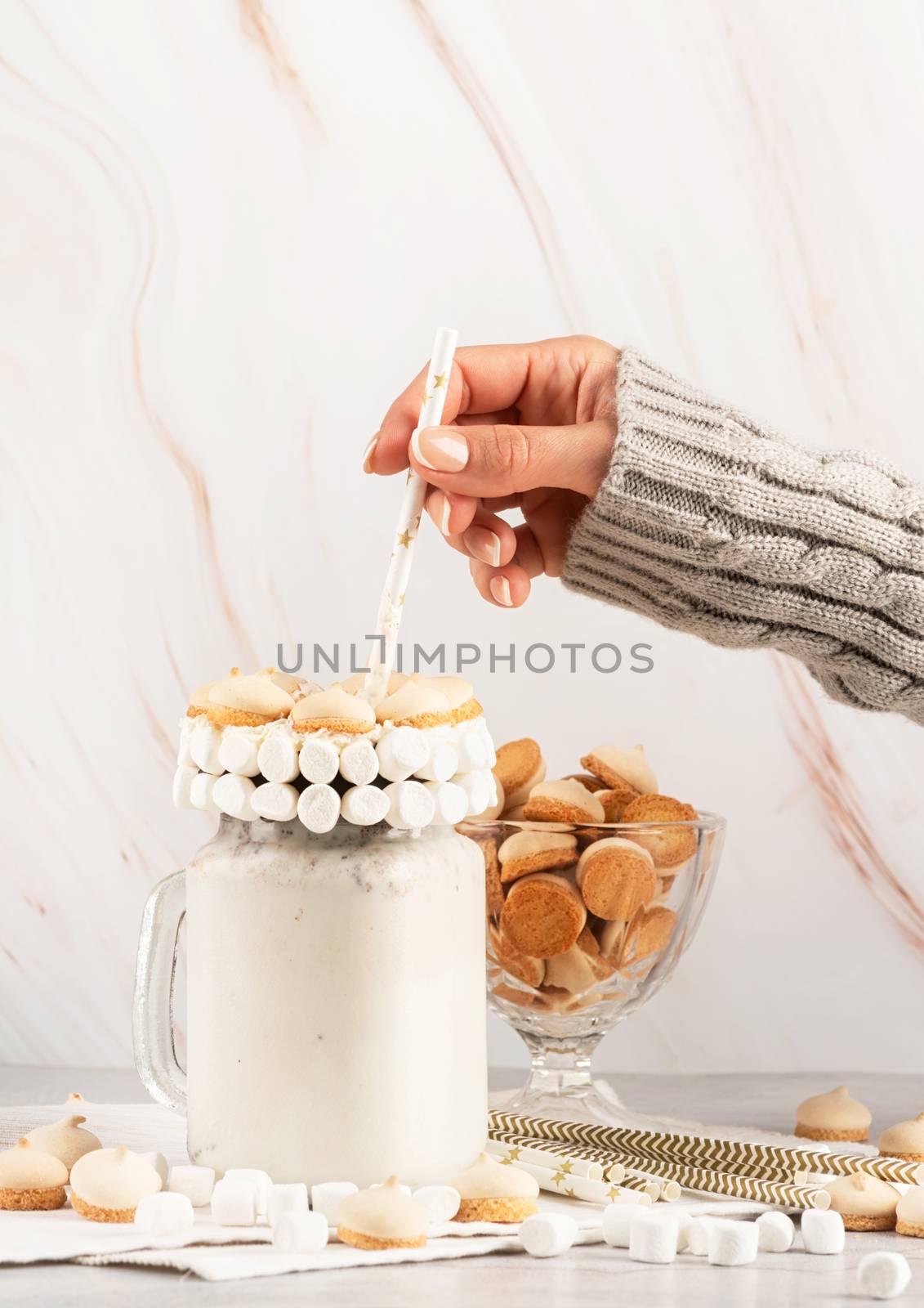 Cold chocolate milkshake in a mason jar decorated with marshamallows and cookies. Woman hand putting a drinking straw into milkshake