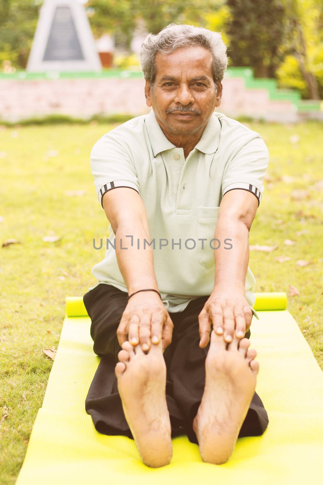 Low-angle full length view of a old man sitting down on exercise or yoga mat touching his toes - Concept of active happy elderly health and fitness by lakshmiprasad.maski@gmai.com