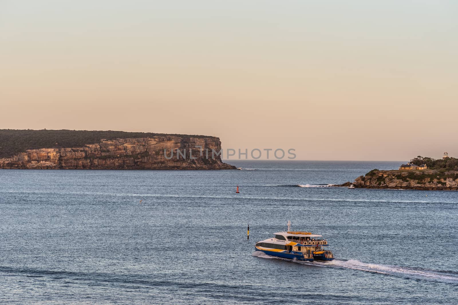 Sydney, Australia - February 12, 2019: North and South Head cliffs at gate between Tasman Sea and Sydney Bay during sunset. Cloudless pale sky. Gray water. Warm brown rocks and a yellow ferry boat.