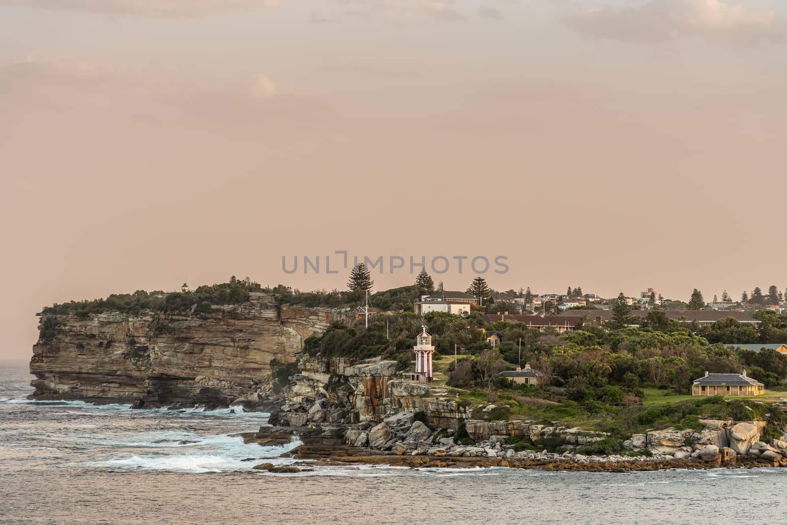 Sydney, Australia - February 12, 2019: South Head cliffs at gate between Tasman Sea and Sydney Bay during sunset. Cloudless pale sky. Gray water. Crashing waves. Warm brown rocks, Hornby lighthouse.