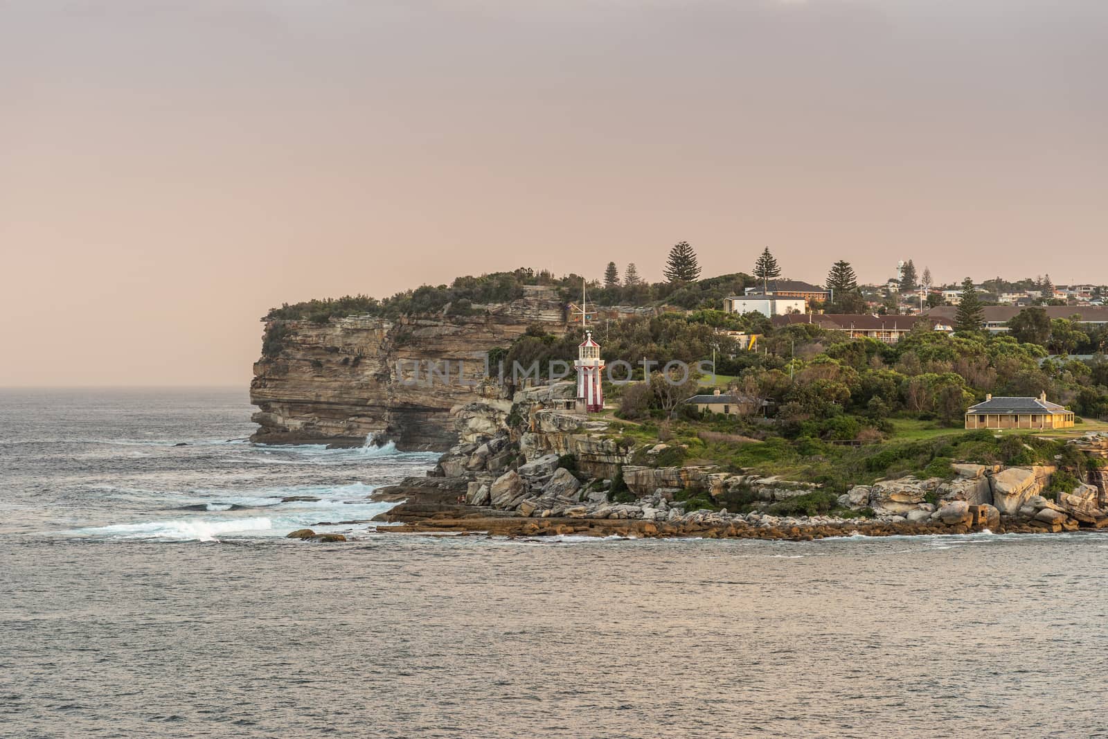 Sydney, Australia - February 12, 2019: South Head cliffs at gate between Tasman Sea and Sydney Bay during sunset. Cloudless pale sky. Gray water. Crashing waves. Warm brown rocks and Hornby lighthouse