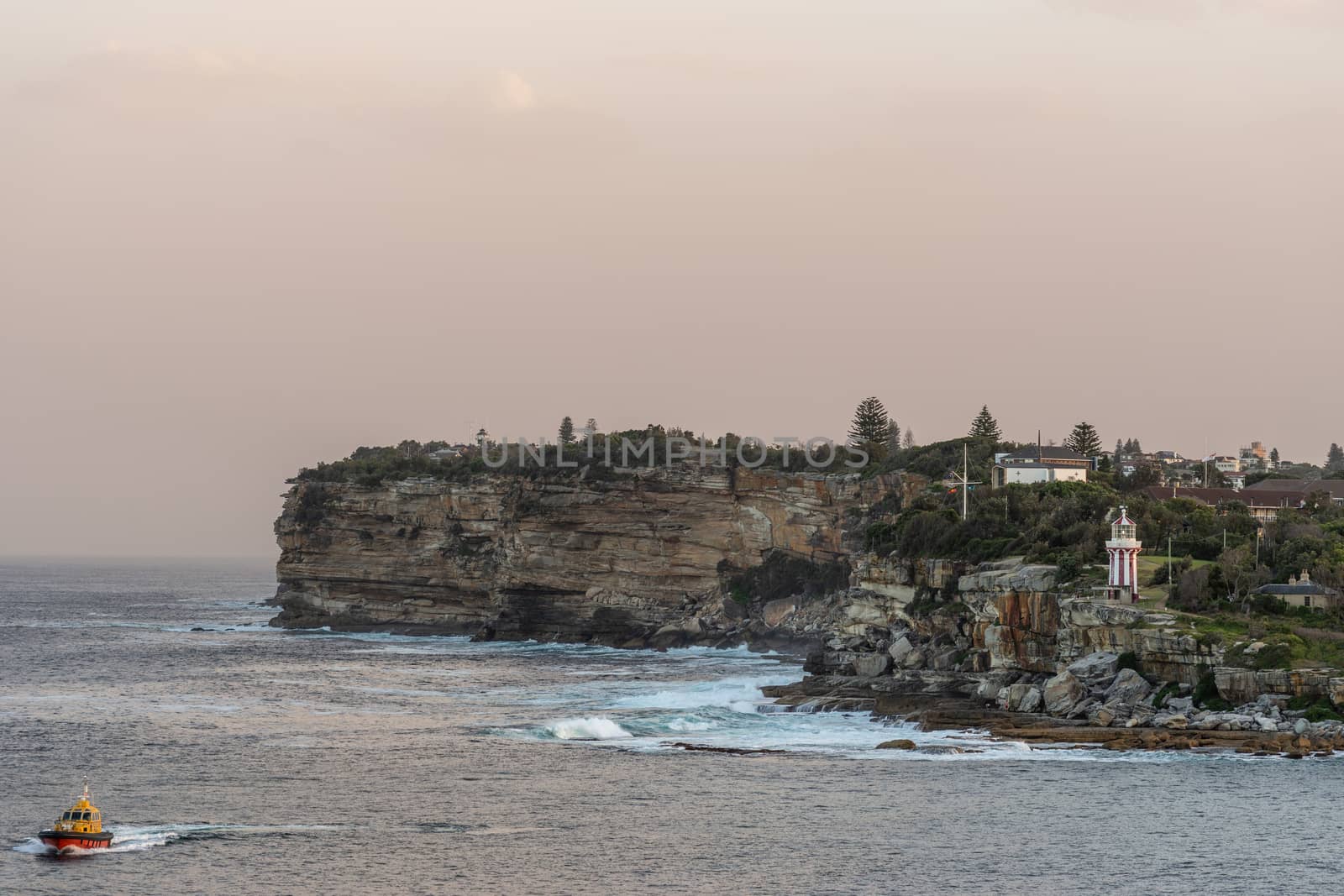 Sydney, Australia - February 12, 2019: South Head cliffs at gate between Tasman Sea and Sydney Bay during sunset. Cloudless pale sky. Gray water. Pilot boat. Crashing waves. Hornby lighthouse