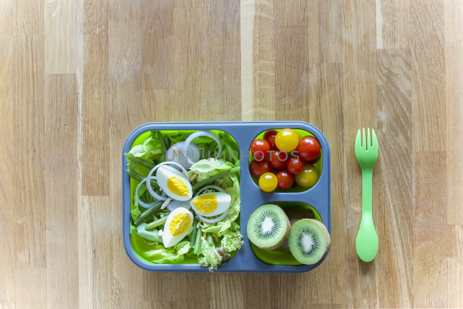 Collapsible silicon lunch box with food (green salads, eggs, tomatoes, kiwi) on wooden table