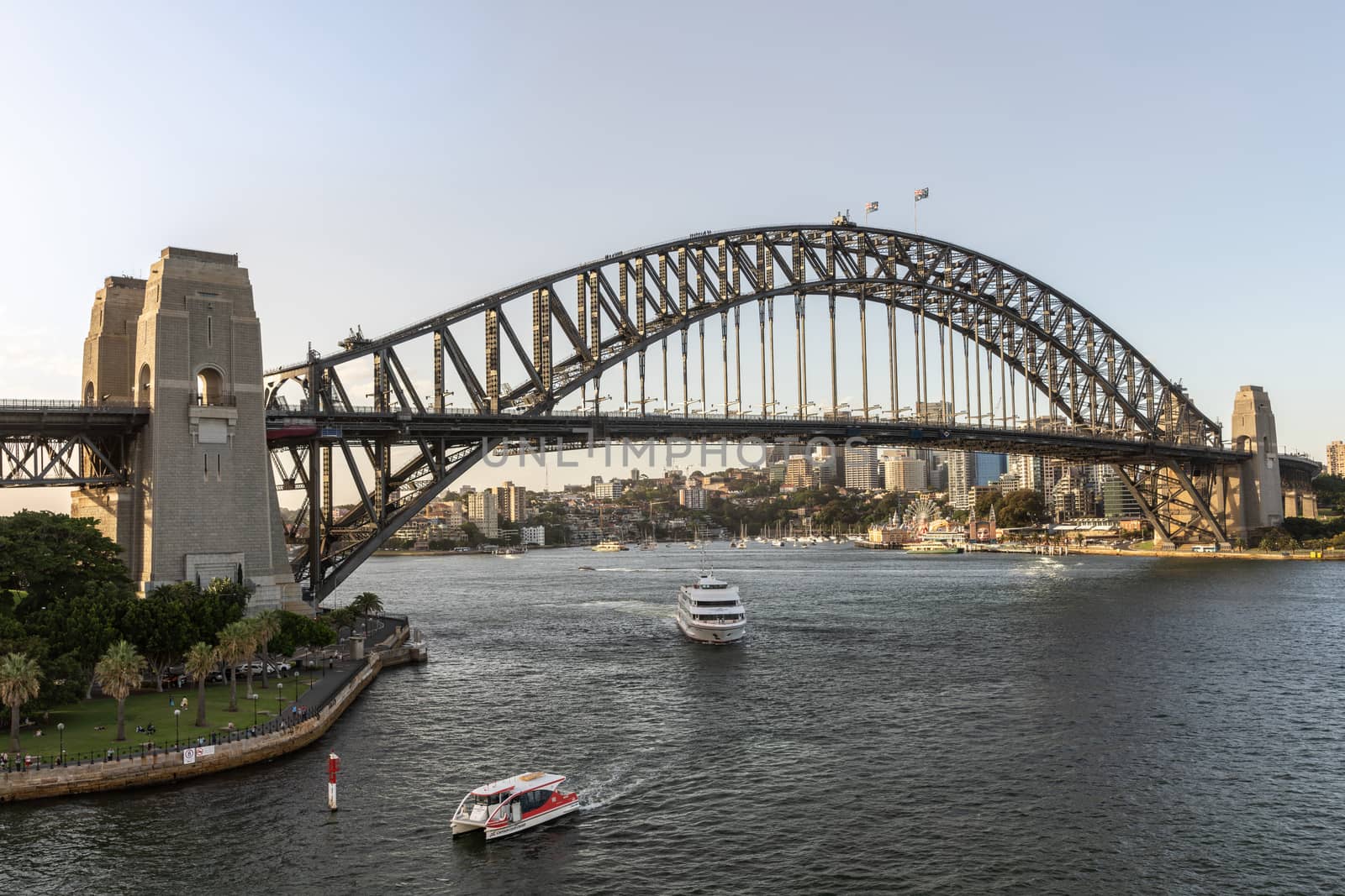 Sydney, Australia - February 12, 2019: Harbour bridge against pale blue sky and boats in the water. Horizon is north shore of bay with Kirribilli neighborhood and Luna Park.