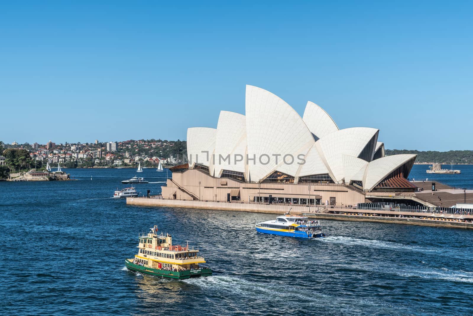 Sydney, Australia - February 12, 2019: Side view of the Opera House with ferries in front. Blue sky and water. Horizon is north shore of bay.