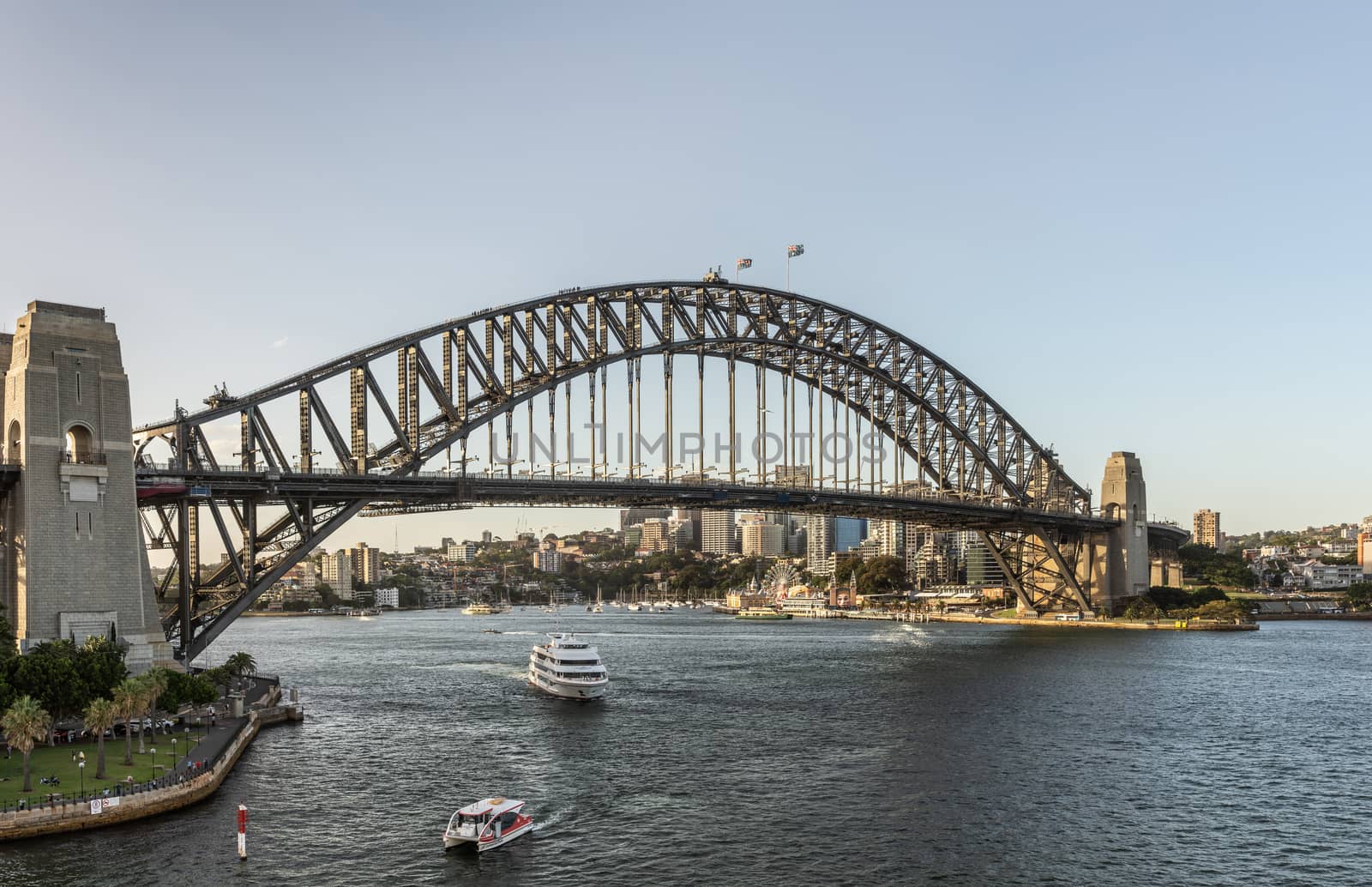 Sydney, Australia - February 12, 2019: Harbour bridge against pale blue sky during sunset. Horizon is north shore of bay with Kirribilli neighborhood and Luna Park. Boats on water.