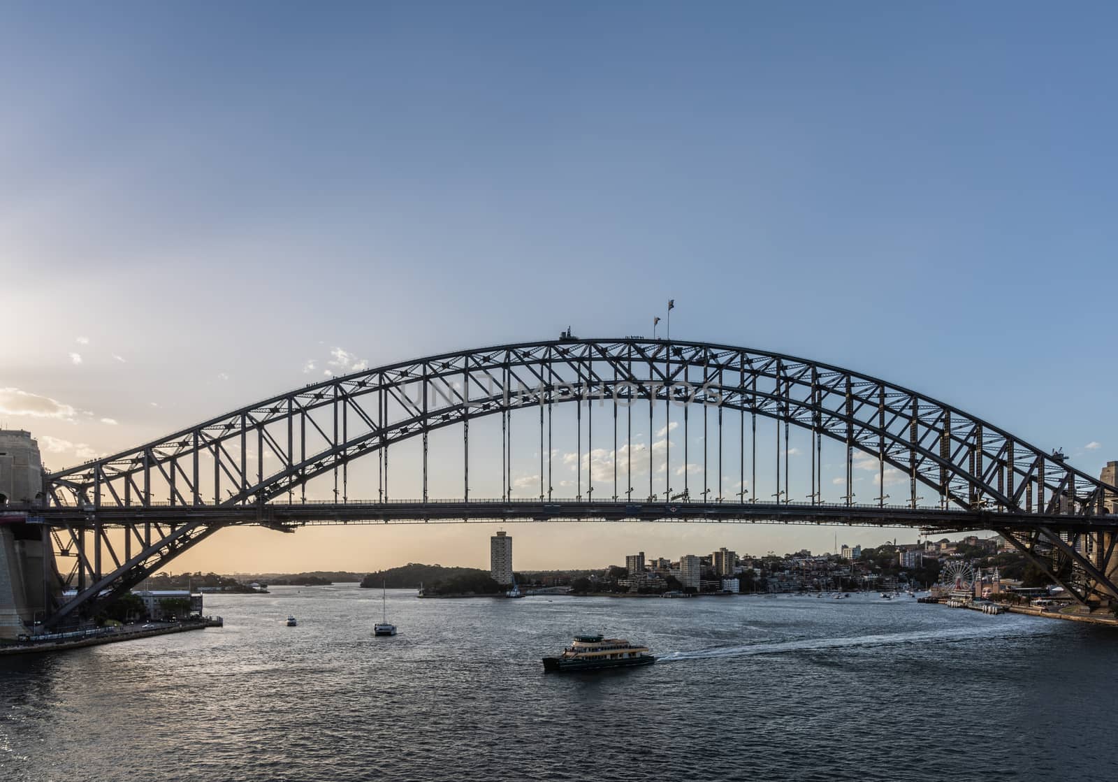 Sydney, Australia - February 12, 2019: Harbour bridge against pale blue sky during sunset. Horizon is north shore of bay with Kirribilli neighborhood and Luna Park. Boats on water.