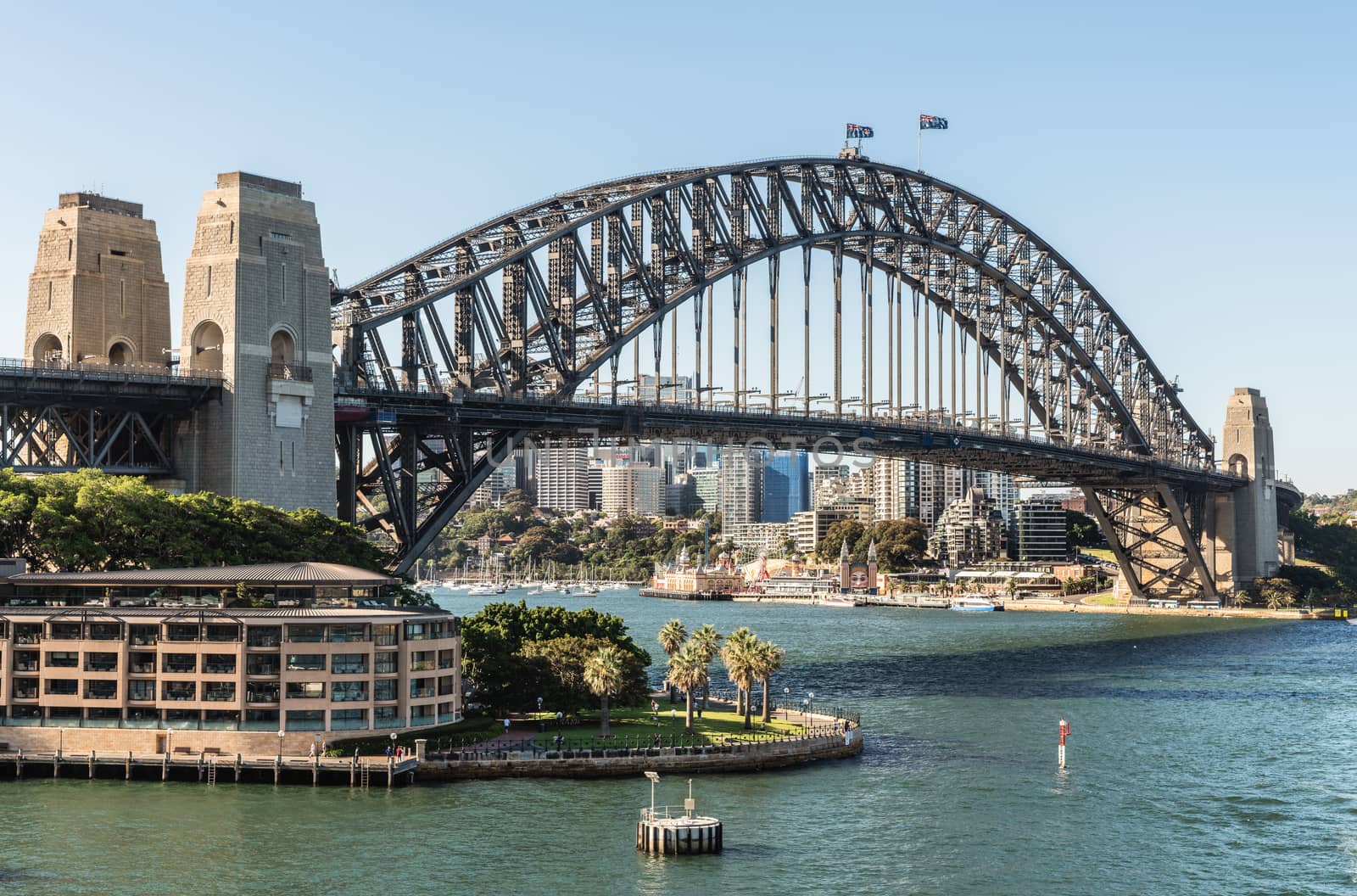 Sydney, Australia - February 12, 2019: Harbour bridge against pale blue sky on late afternoon. Horizon is north shore of bay with Kirribilli neighborhood and Luna Park.