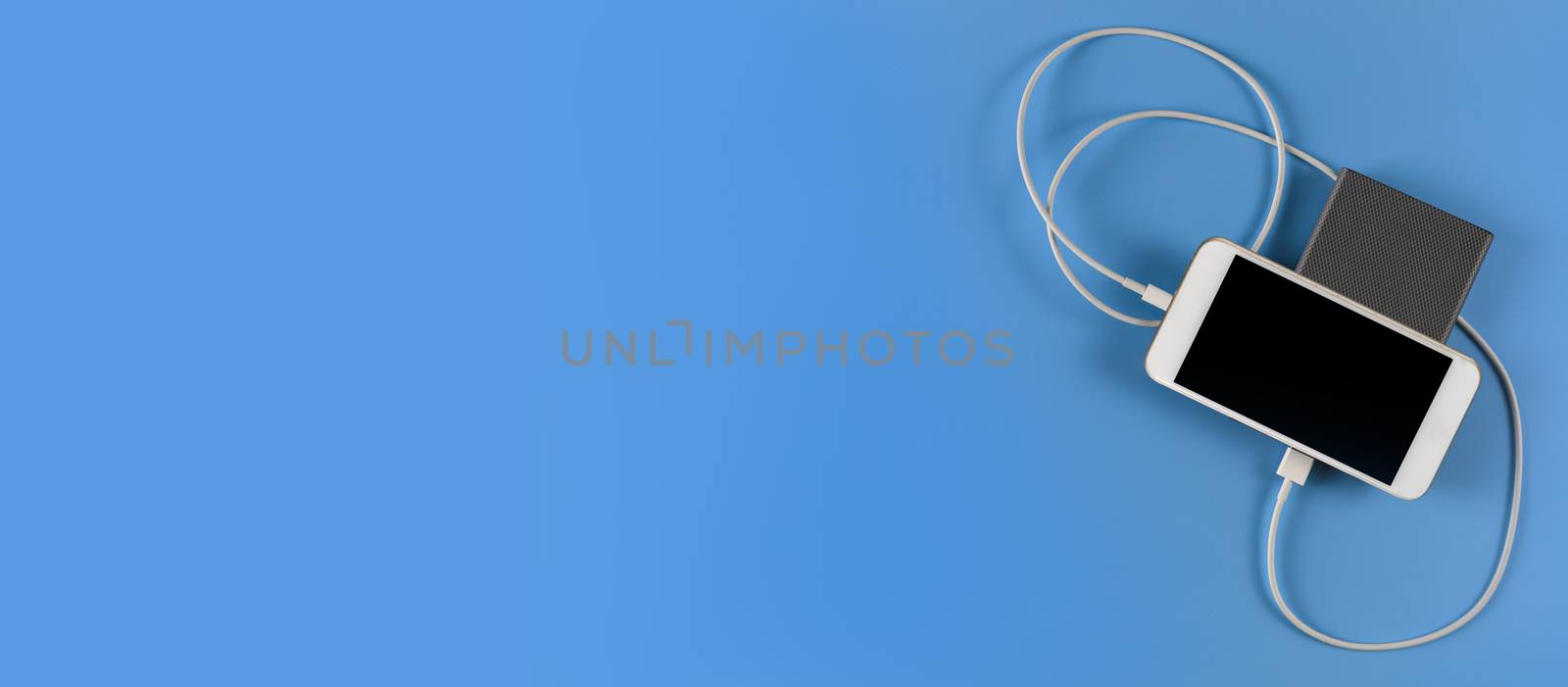 Top view smartphone charging with power bank on blue background with copy space. Smartphone charging with power bank by white USB cable.