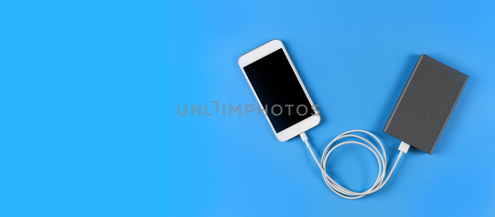 Top view smartphone charging with power bank on blue background with copy space. Smartphone charging with power bank by white USB cable.