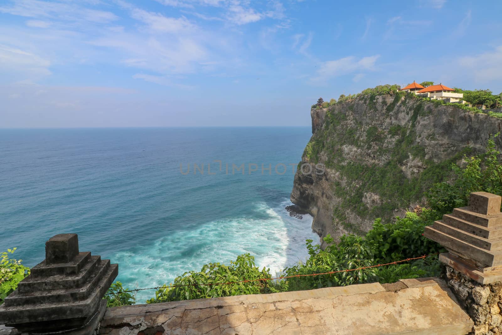View of Uluwatu cliff with pavilion and blue sea in Bali, Indonesia. Beautiful scenery of Pura Luhur Uluwatu Temple with colorful flowers foreground during day. Scenic coastal landscape of high cliff.