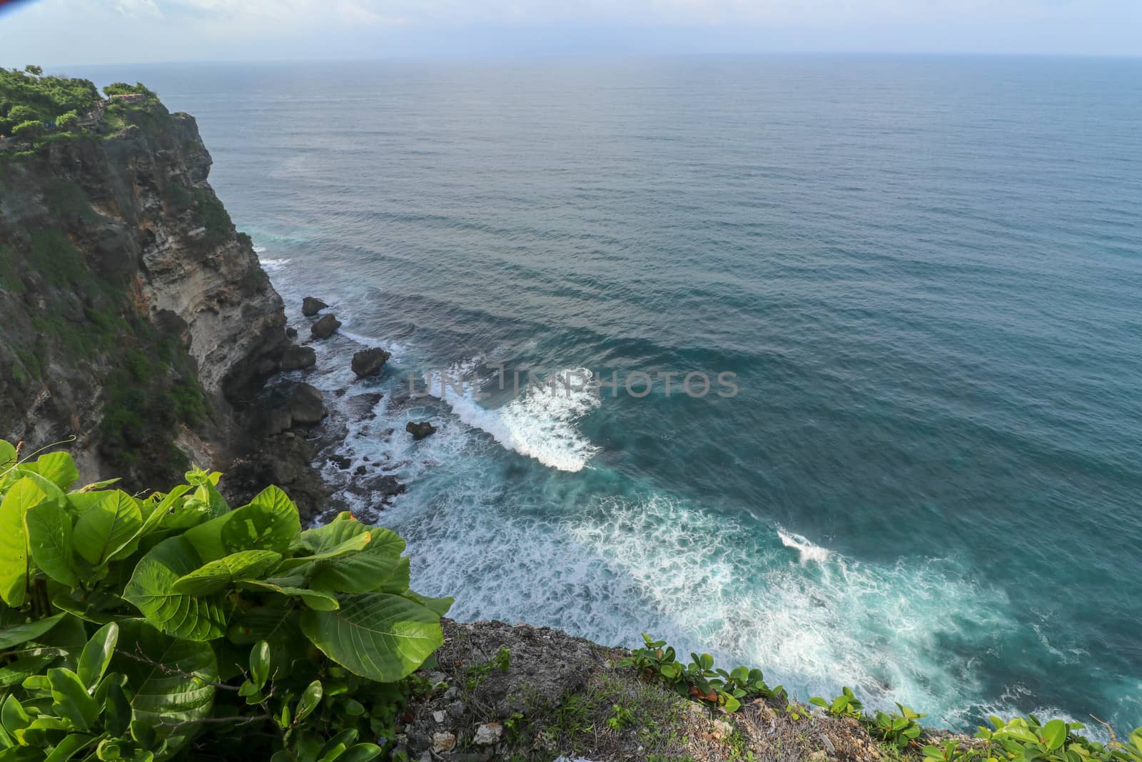 View of Uluwatu cliff with pavilion and blue sea in Bali, Indonesia. Beautiful scenery of Pura Luhur Uluwatu Temple with colorful flowers foreground during day. Scenic coastal landscape of high cliff.