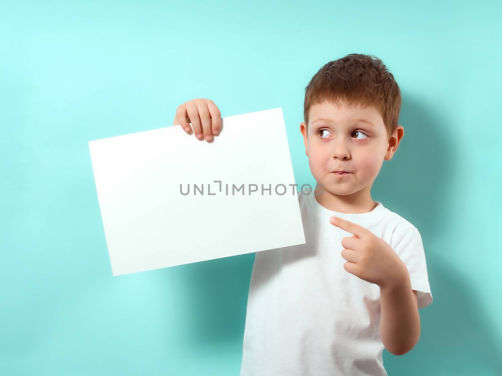 Four-year-old boy with a surprised face, looking away and shows finger on blank white sheet. Fun child on blue background with copy space for message, mock up