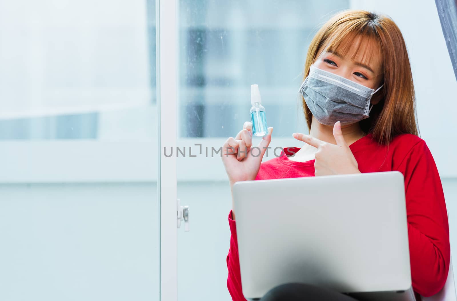 Work from home concept, Asian beautiful young woman smiling, student girl sitting on chair during working with laptop computer at home office and pointing alcohol spray sanitizing on hand