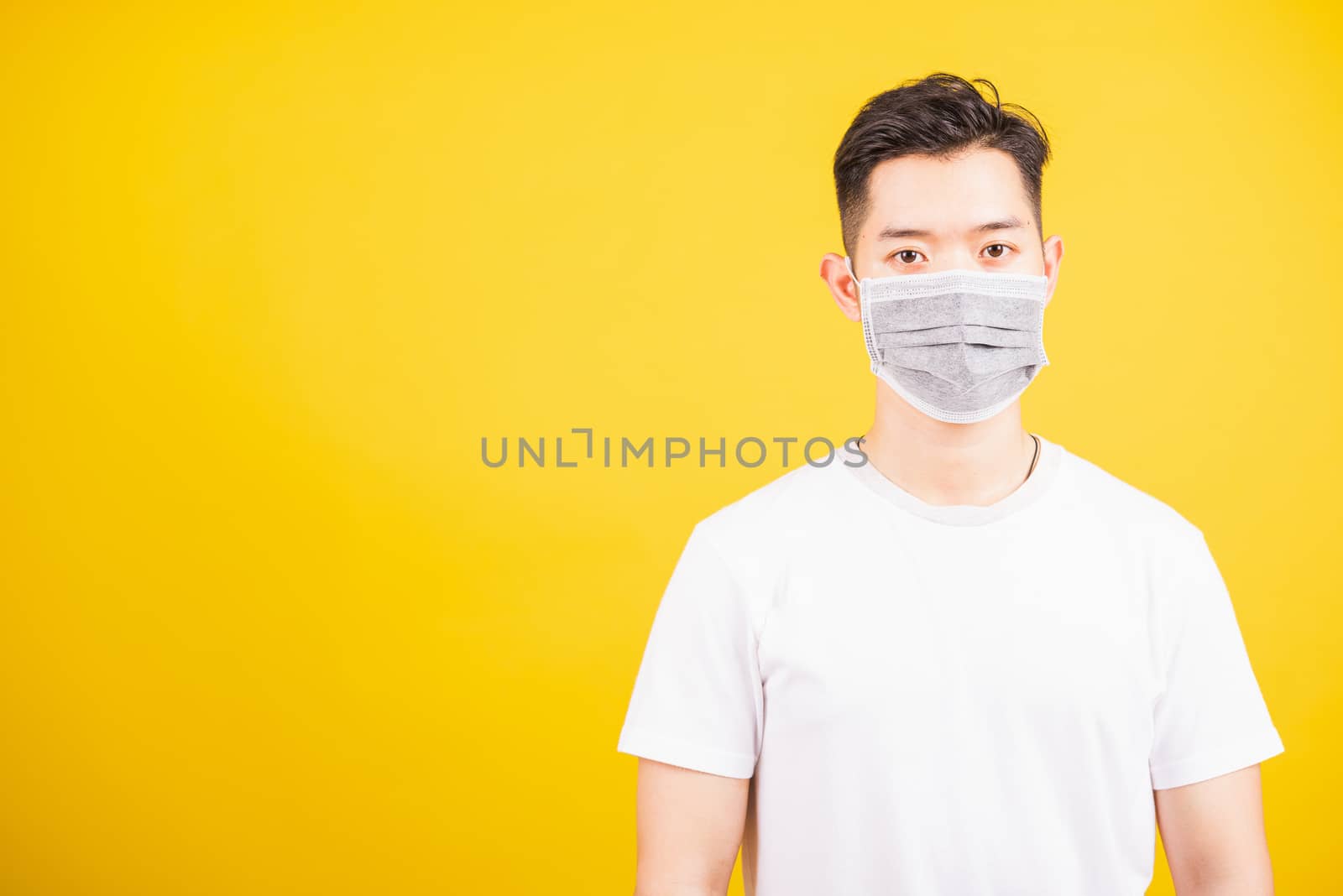 Closeup Asian young man wearing face mask protective germ virus or air dust, studio shot isolated on yellow background and copy space, medical outbreak coronavirus COVID-19 concept