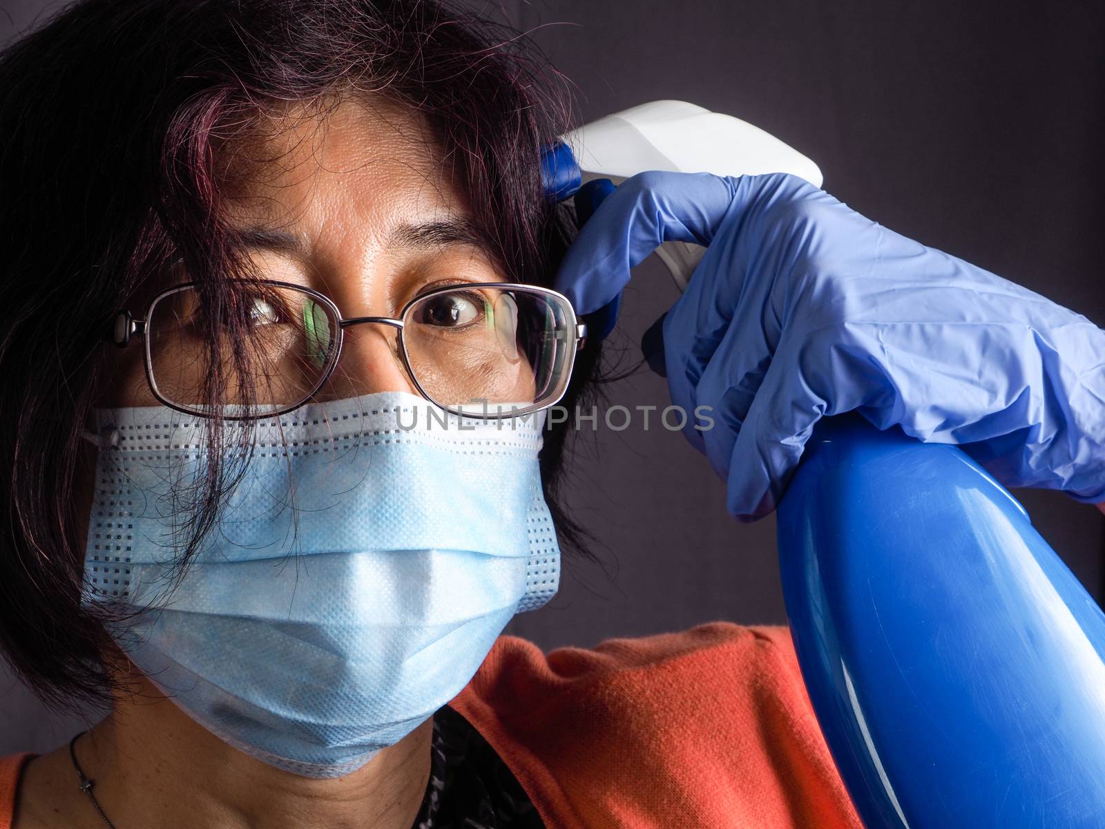 funny humorous adult asian 40s 50s woman wearing protection surgical face mask , blue latex gloves, and sun glasses pointing the head with blue sanitizer cleaning  bottle as a weapon as tired of confinement covid concept.