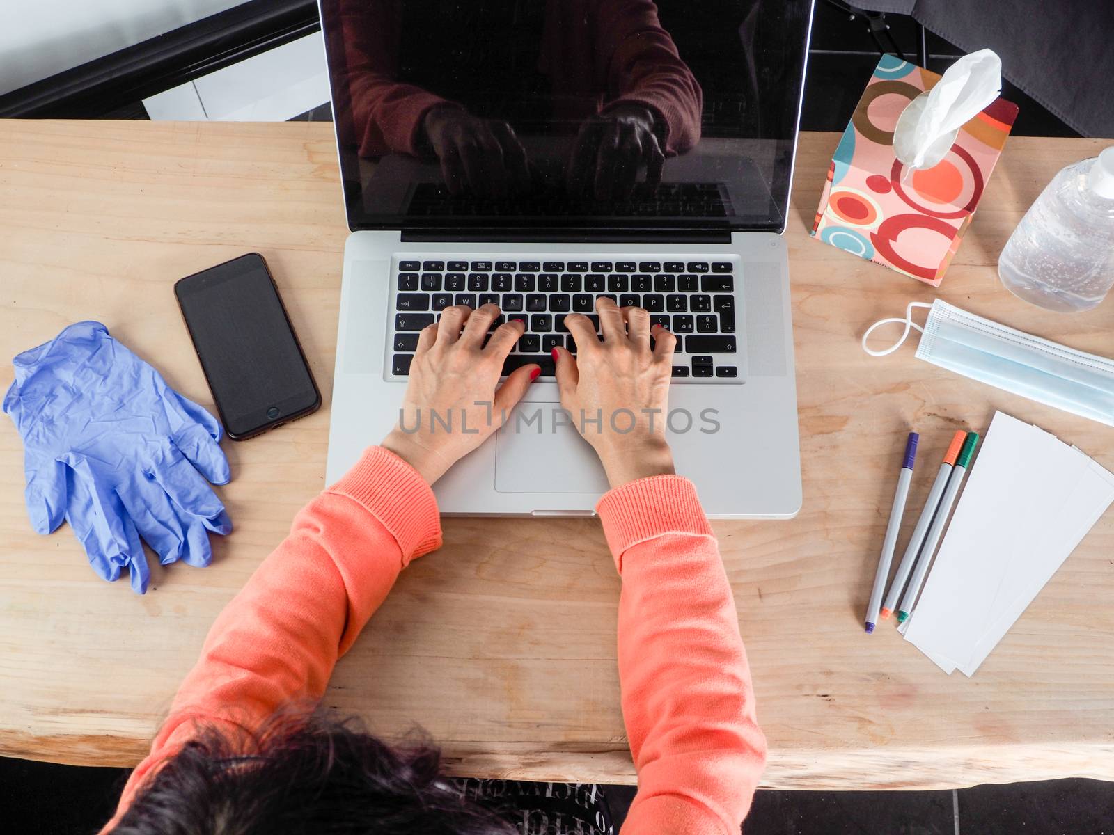 Woman hand s freelance working on a notebook laptop computer at home or home office with gel and face mask prevention items near connected to internet via hot spot of smartphone in fall, orange sweater and brown table, earth tones.