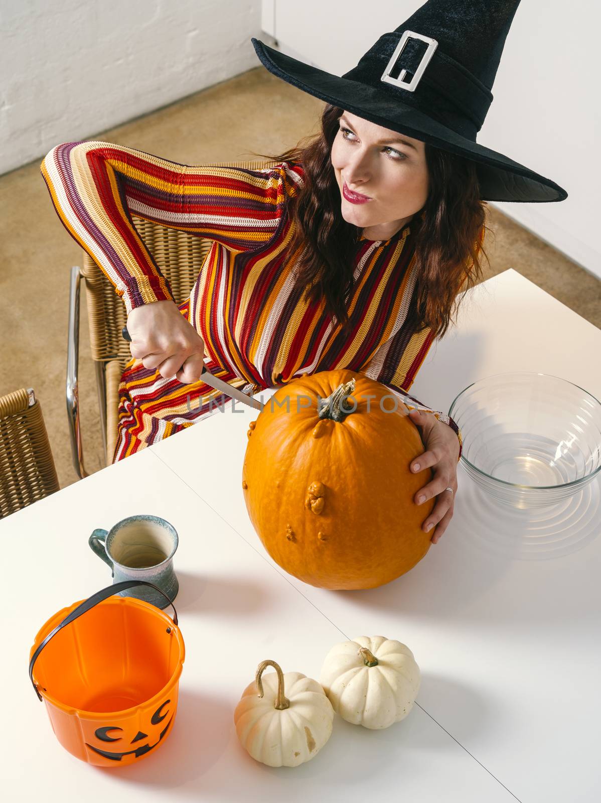 Beautiful young woman wearing witch's black hat in her kitchen cutting a pumpkin for Halloween.