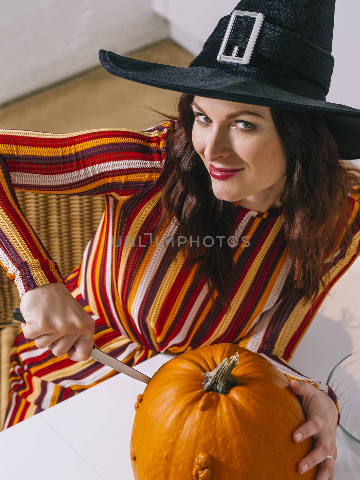 Beautiful young woman wearing witch's black hat  in her kitchen cutting a pumpkin for Halloween.