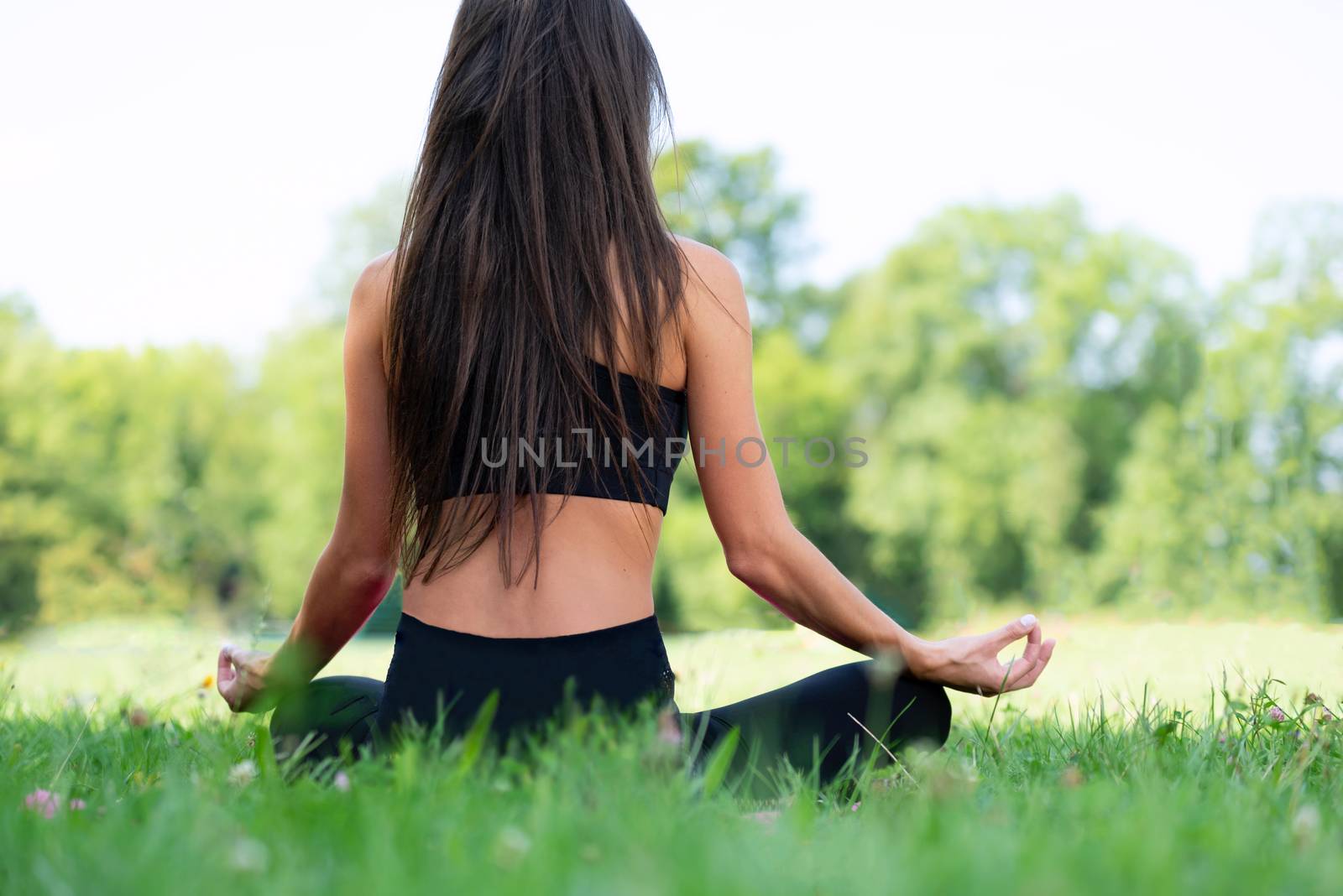 Back view of a young woman doing yoga meditation in a lotus pose outdoors in a park on a sunny day