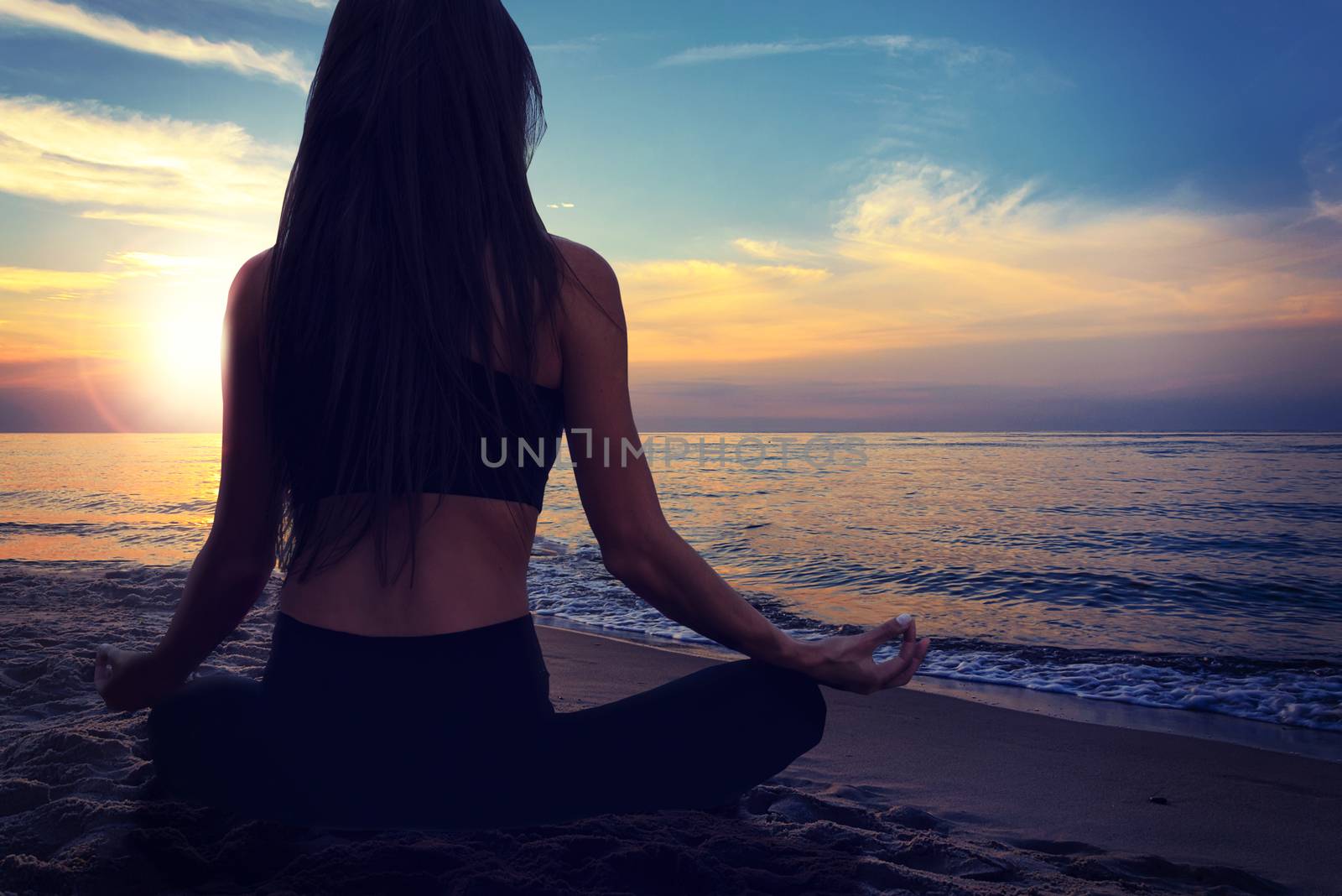 Back view of a young woman doing yoga meditation in a lotus pose at the seacoast on a sunset background.