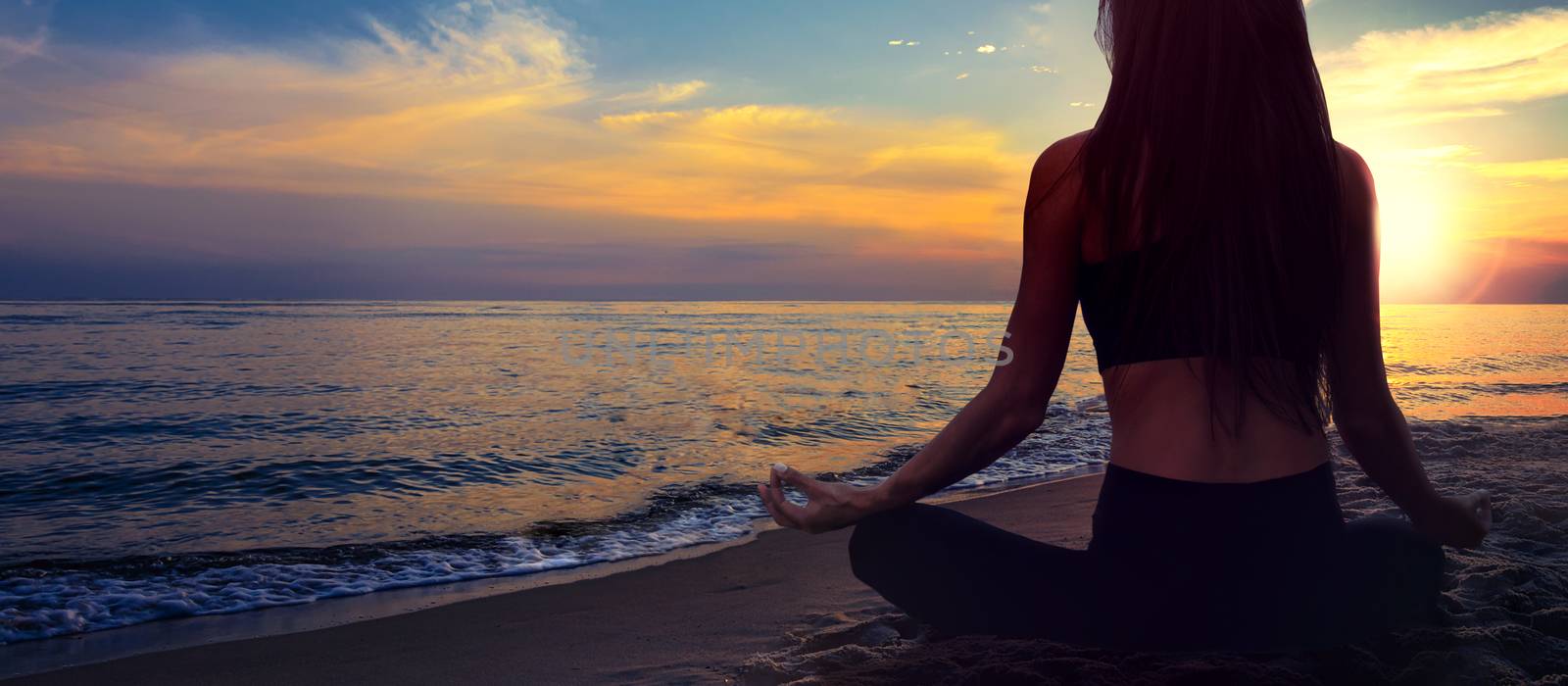 Yoga meditation banner - woman in a lotus pose on a seaside beach on a background of a picturesque sunset landscape (copy space)