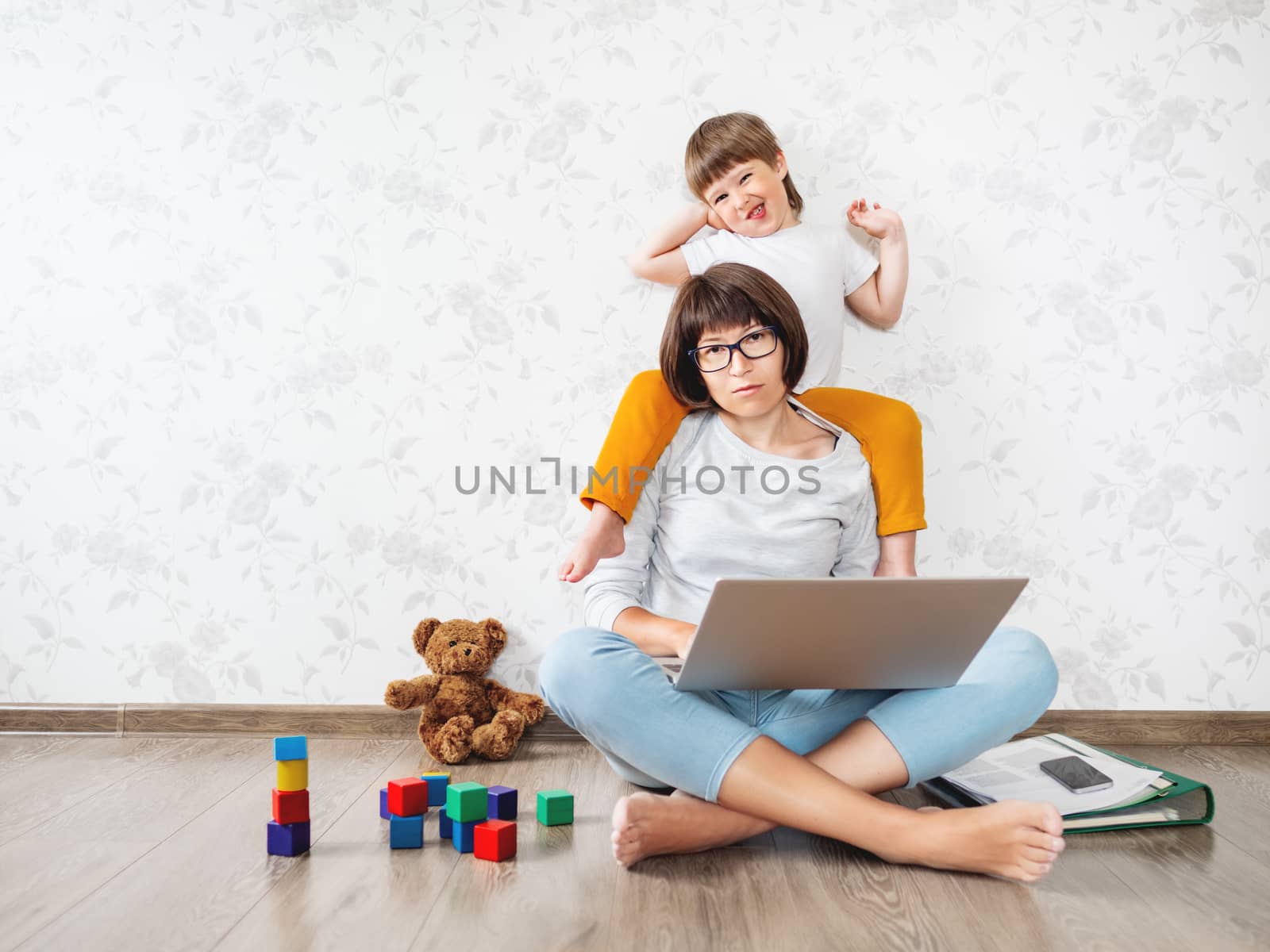 Mom and son at home. Mother works remotely with laptop, son sits on her neck. Freelance work in same time with raising children.