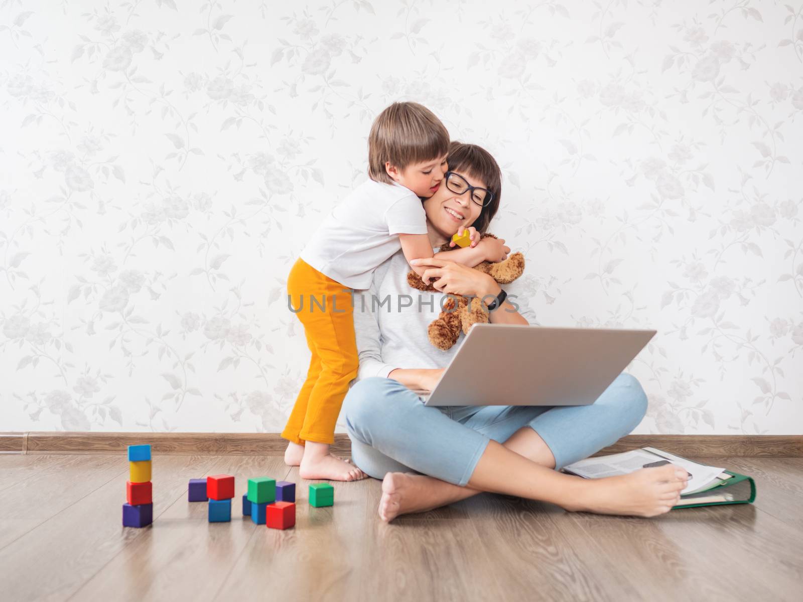 Mom and son at home. Mother works remotely with laptop, son hugs her. Freelance work in same time with raising children.