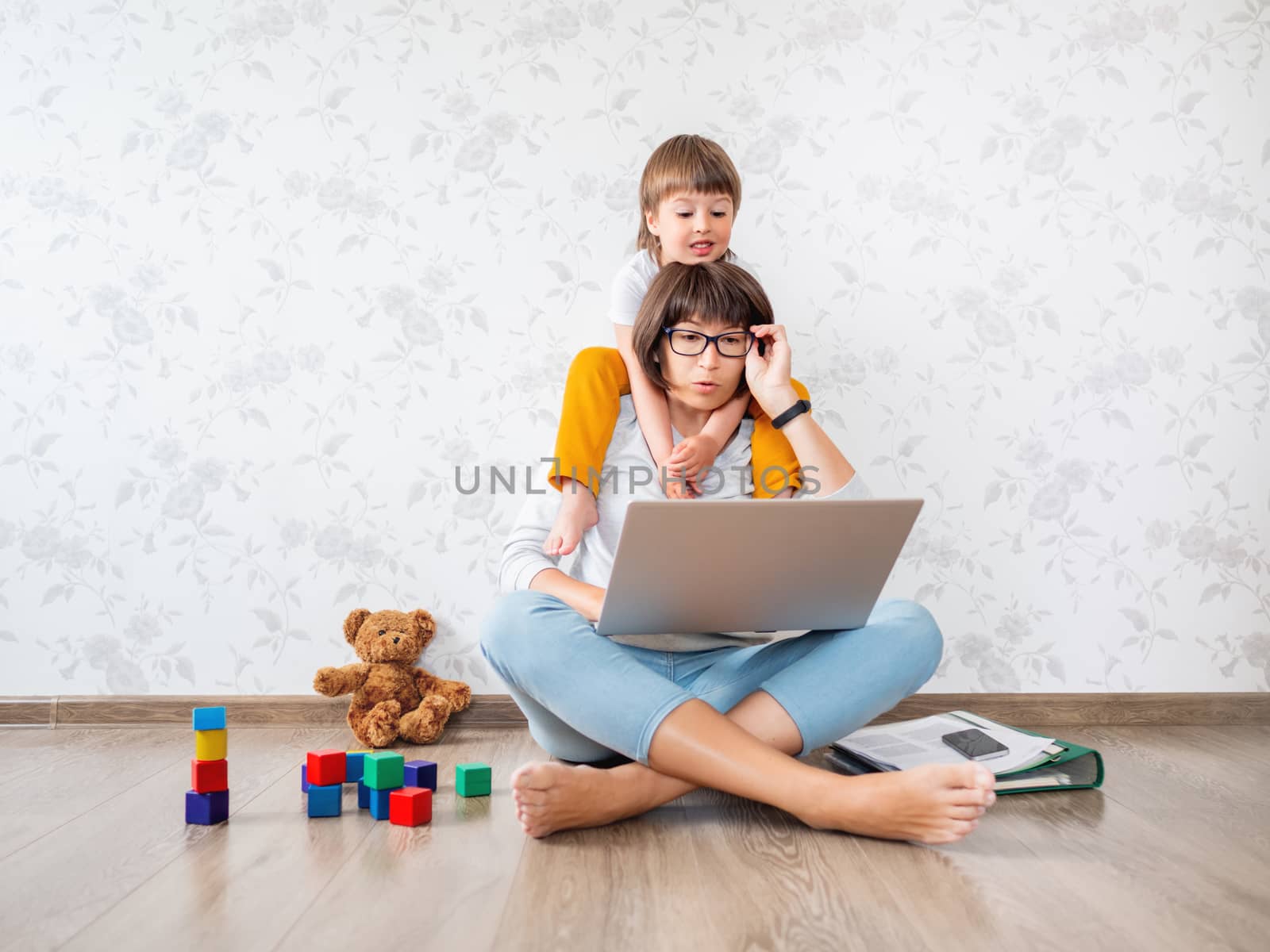 Mom and son at home. Mother works remotely with laptop, son sits on her neck. Freelance work in same time with raising children.
