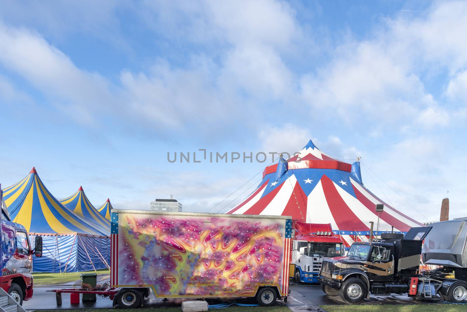 Circus caravanes in front the red and white circus tents topped with bleu starred cover against a sunny blue sky with clouds by ankorlight