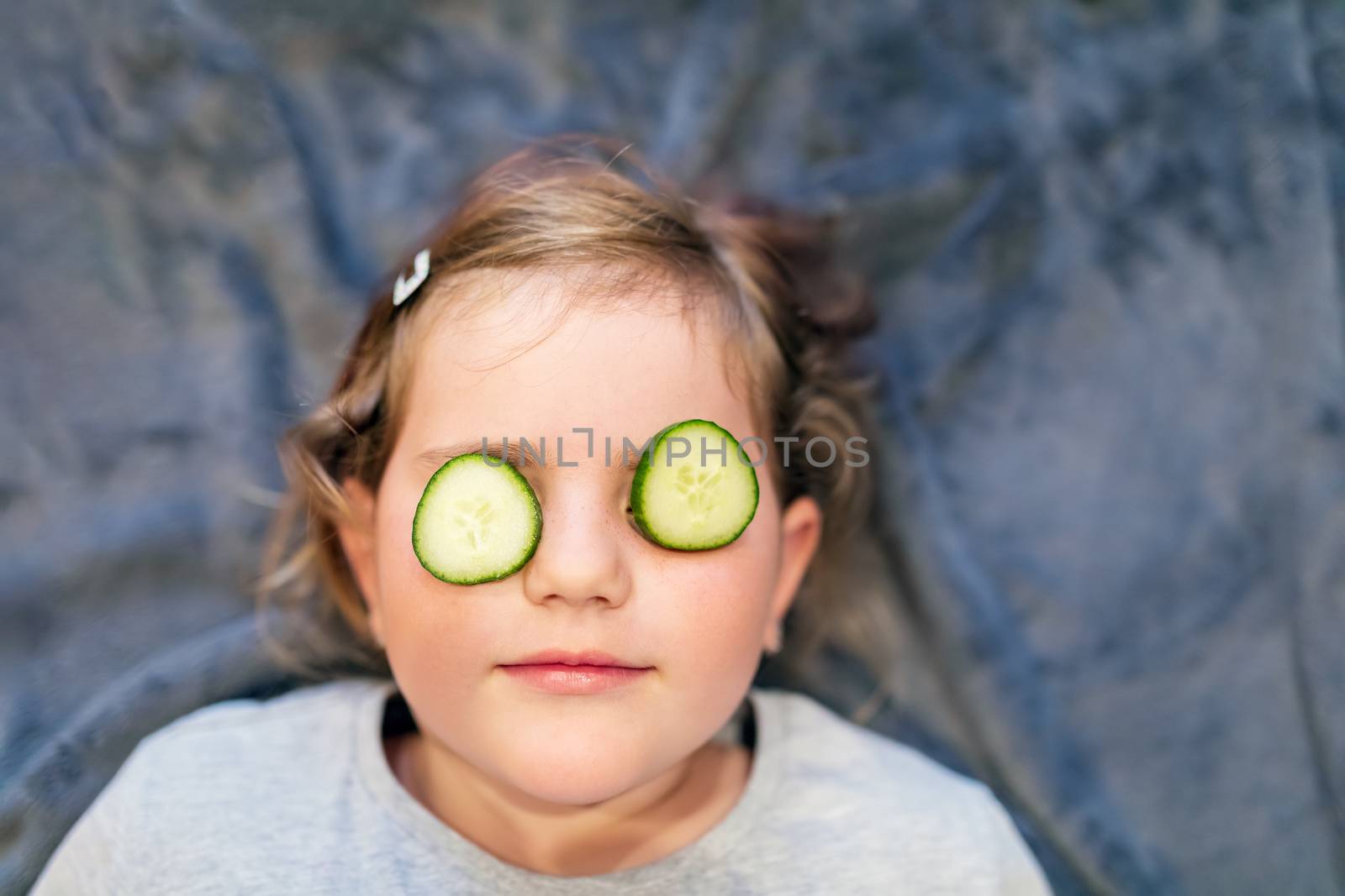 Funny small girl with piece of cucumber on their eyes like a mask, beauty and health concept, indoor closeup portrait