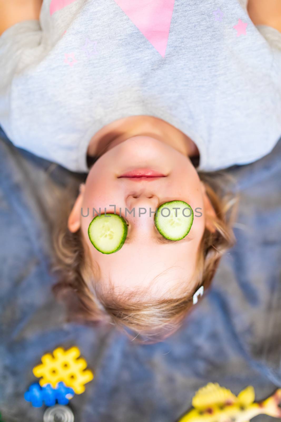 Funny small girl with piece of cucumber on their eyes like a mask, beauty and health concept, indoor closeup portrait