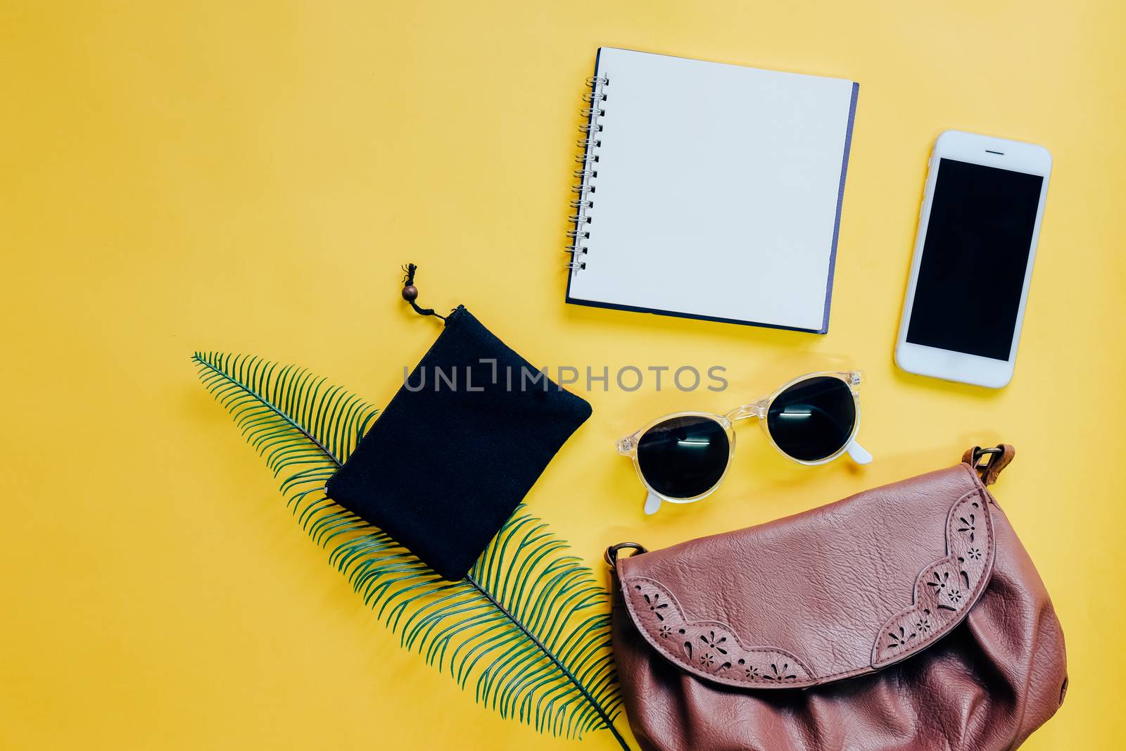 Top view flat lay of woman bag with smartphone, blank notebook, sunglasses and green leaf on yellow background, copy space