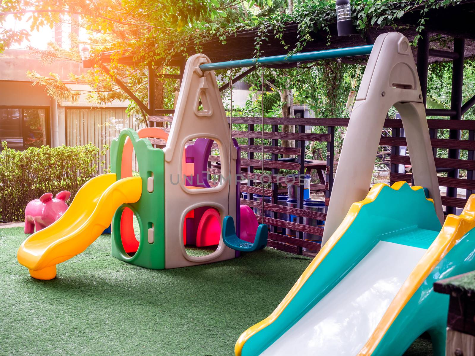 Colorful playground equipment on green yard, small area for children. Swing seats and garden slides on green grass with warm sunlight in the morning.