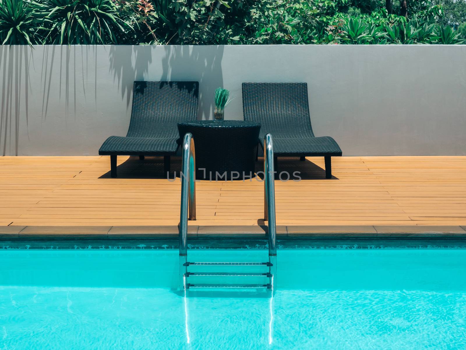 Outdoor swimming pool background minimal style. Grab bars ladder in the blue swimming pool with two black sunbed and green shrubs on grey wall.