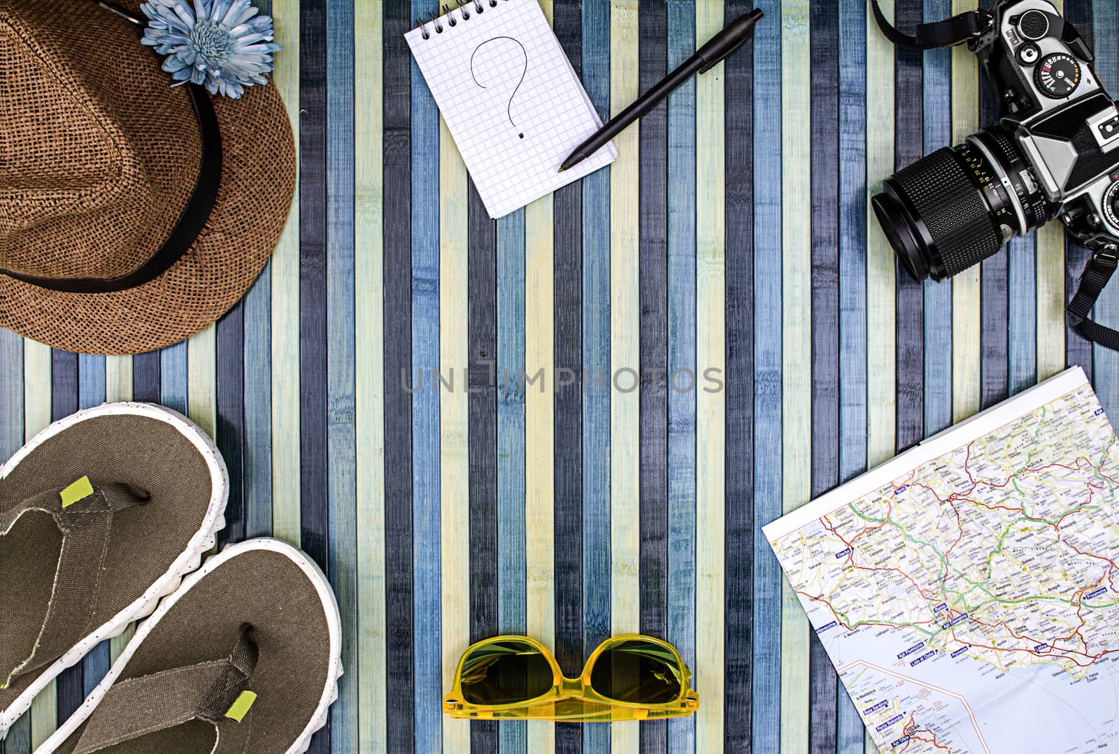 Background composition from above for summer holidays with vintage camera, sunglasses, flip-flops, straw hat, map and notepad with handwritten question mark. Striped wooden background on the light blue.