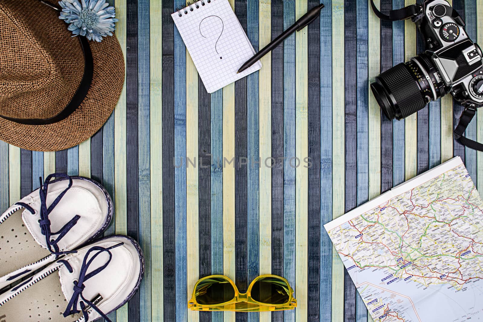 Background composition from above for summer holidays with vintage camera, sunglasses, white shoes, straw hat, map and notepad with handwritten question mark. Striped wooden background on the light blue.
