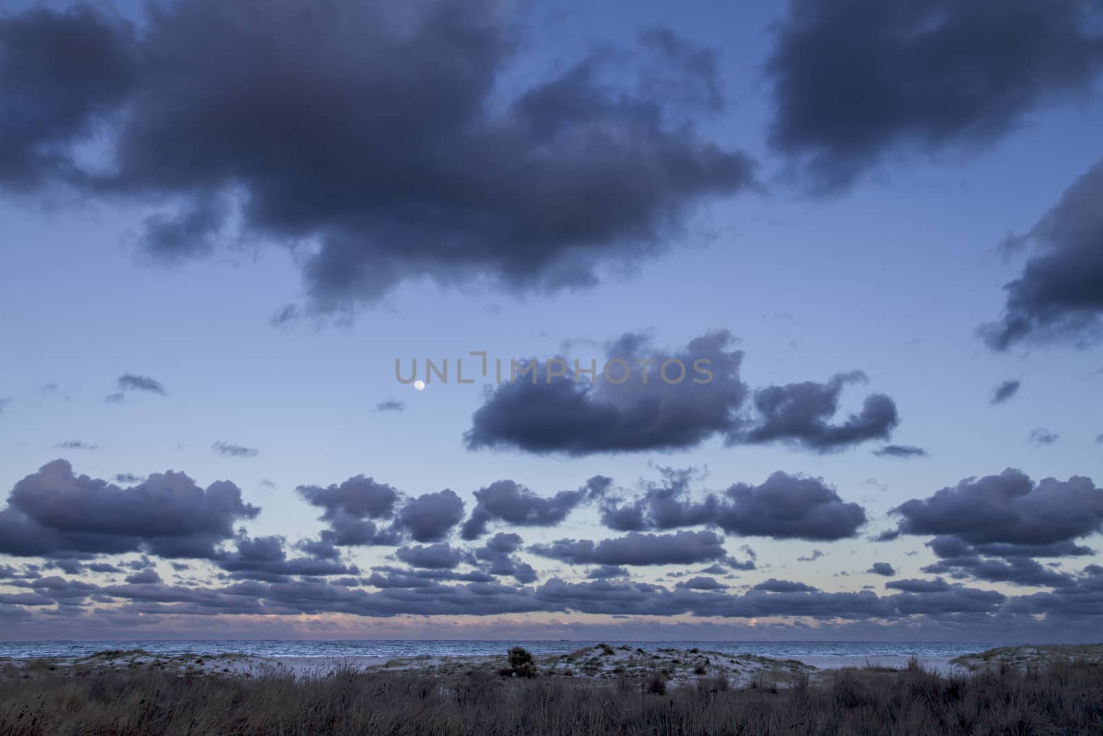 Impressive sunset sky with the full moon and layers of clouds that slope down towards the horizon over the dark sea and the white beach