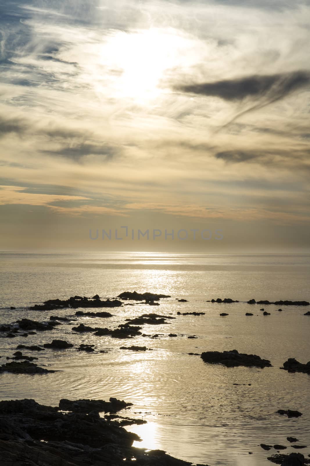 Blue sky at dawn covered by strange cloud formations on a flat sea with black rocks that stand out in the reflection of the sun on the water