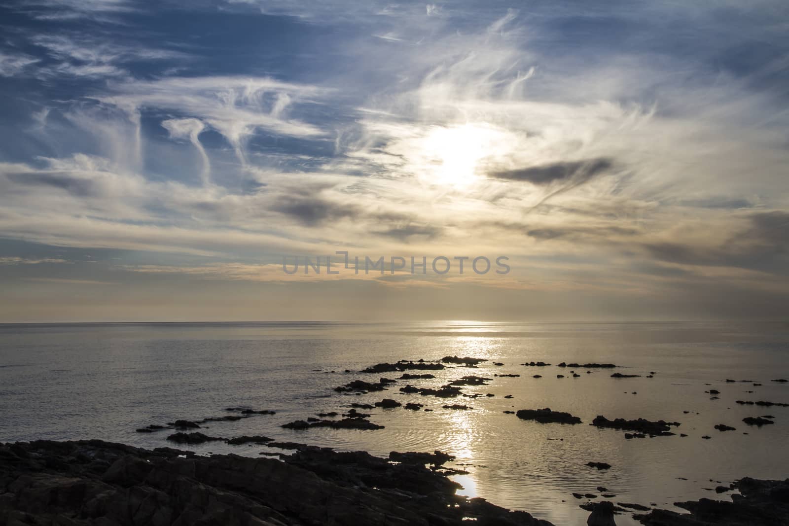 Blue sky at dawn covered by strange cloud formations on a flat sea with black rocks that stand out in the reflection of the sun on the water