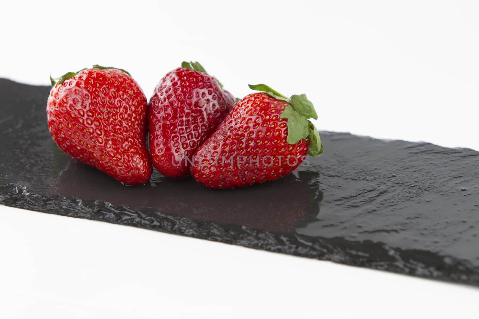 Close-up of three isolated strawberries on a rectangular strip of diagonally wet slate on a white background shot in high angle view 