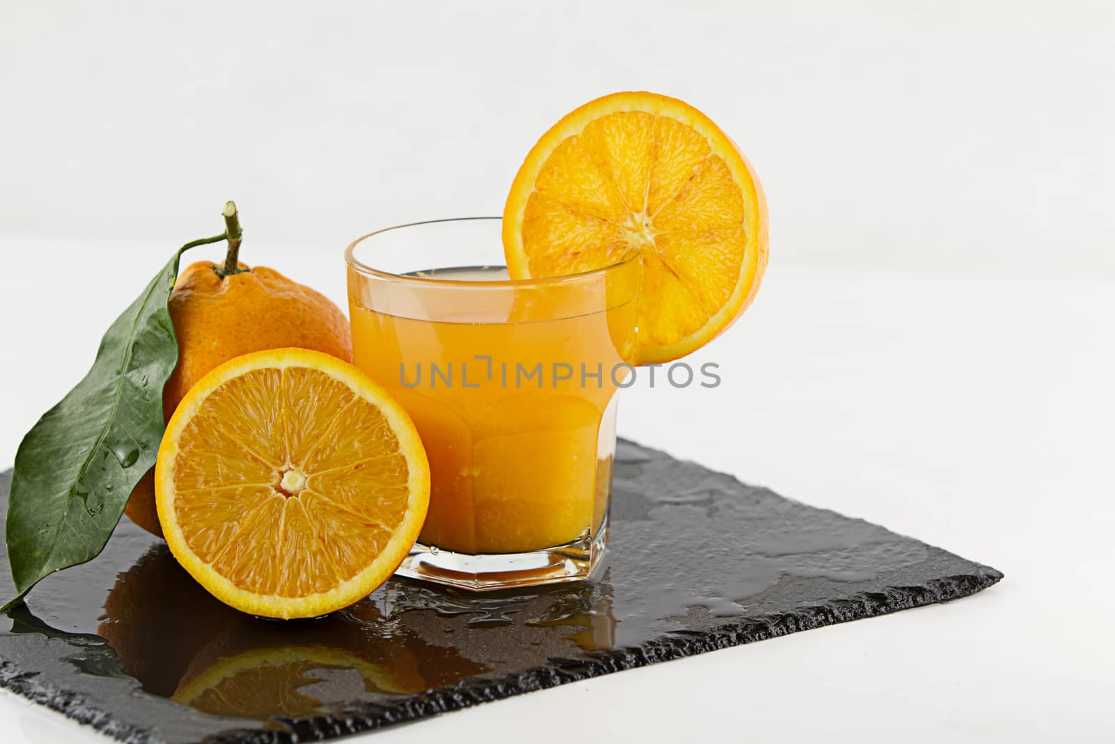 An inviting glass full of orange juice with orange slice on the rim, a half orange and a whole one with leaf on a square wet slate plate on white background