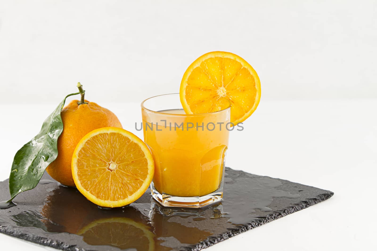 An inviting glass full of orange juice with orange slice on the rim, a half orange and a whole one with leaf on a square wet slate plate on white background