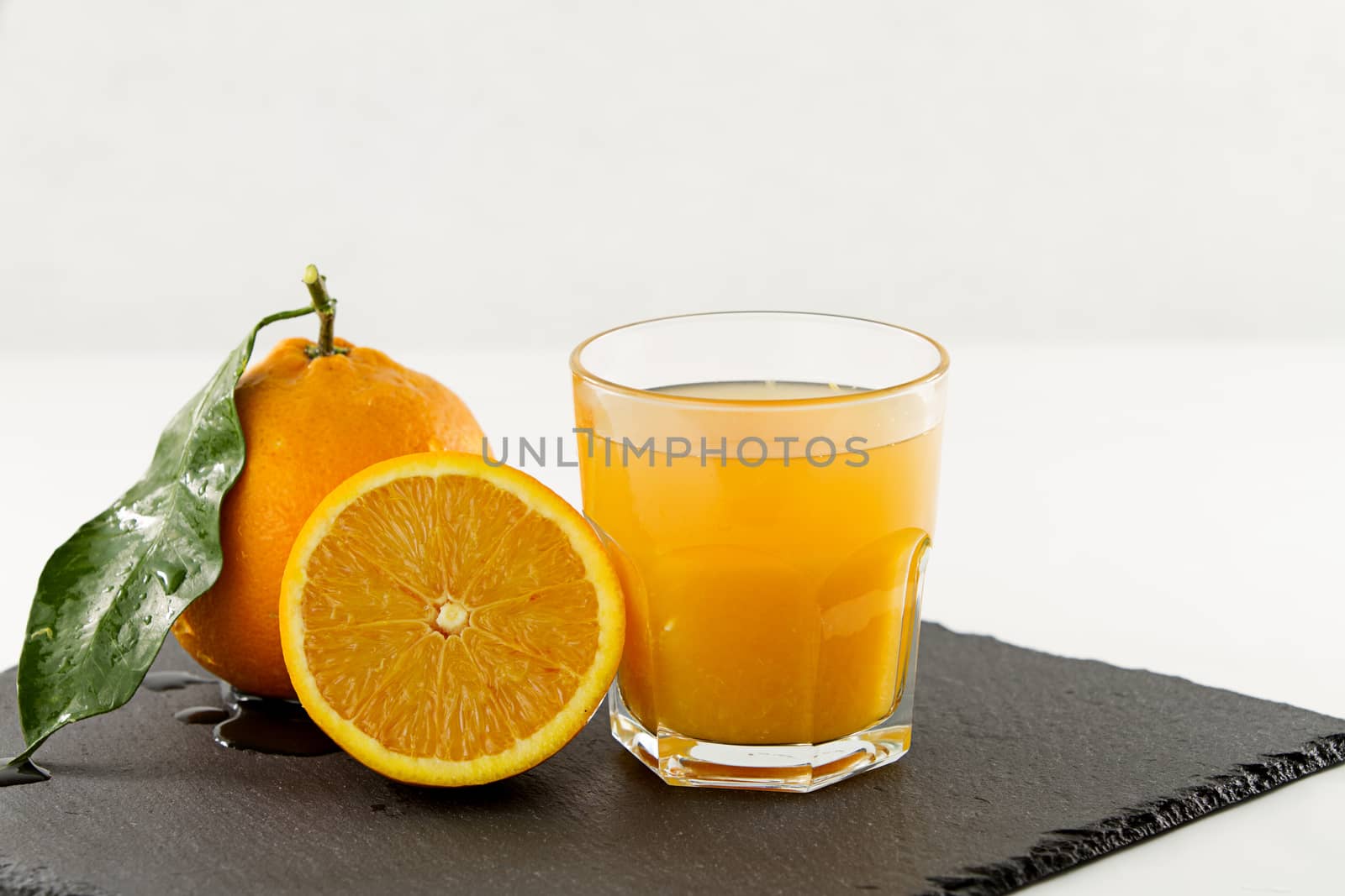 An inviting glass full of orange juice, a half orange and a whole one with leaf on a square slate plate onwhite background